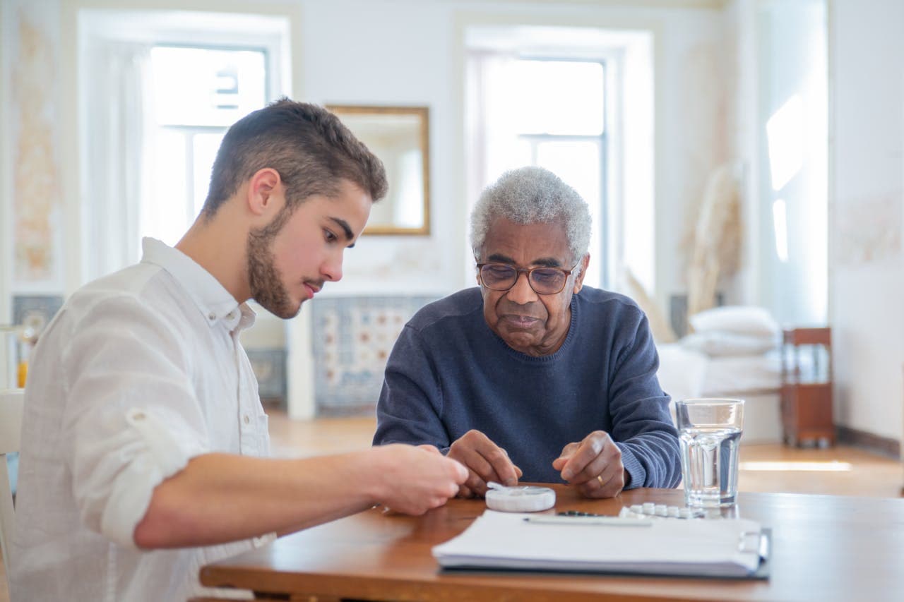 A young man sitting at a table with an elderly man, dividing up a weekly dose of medications